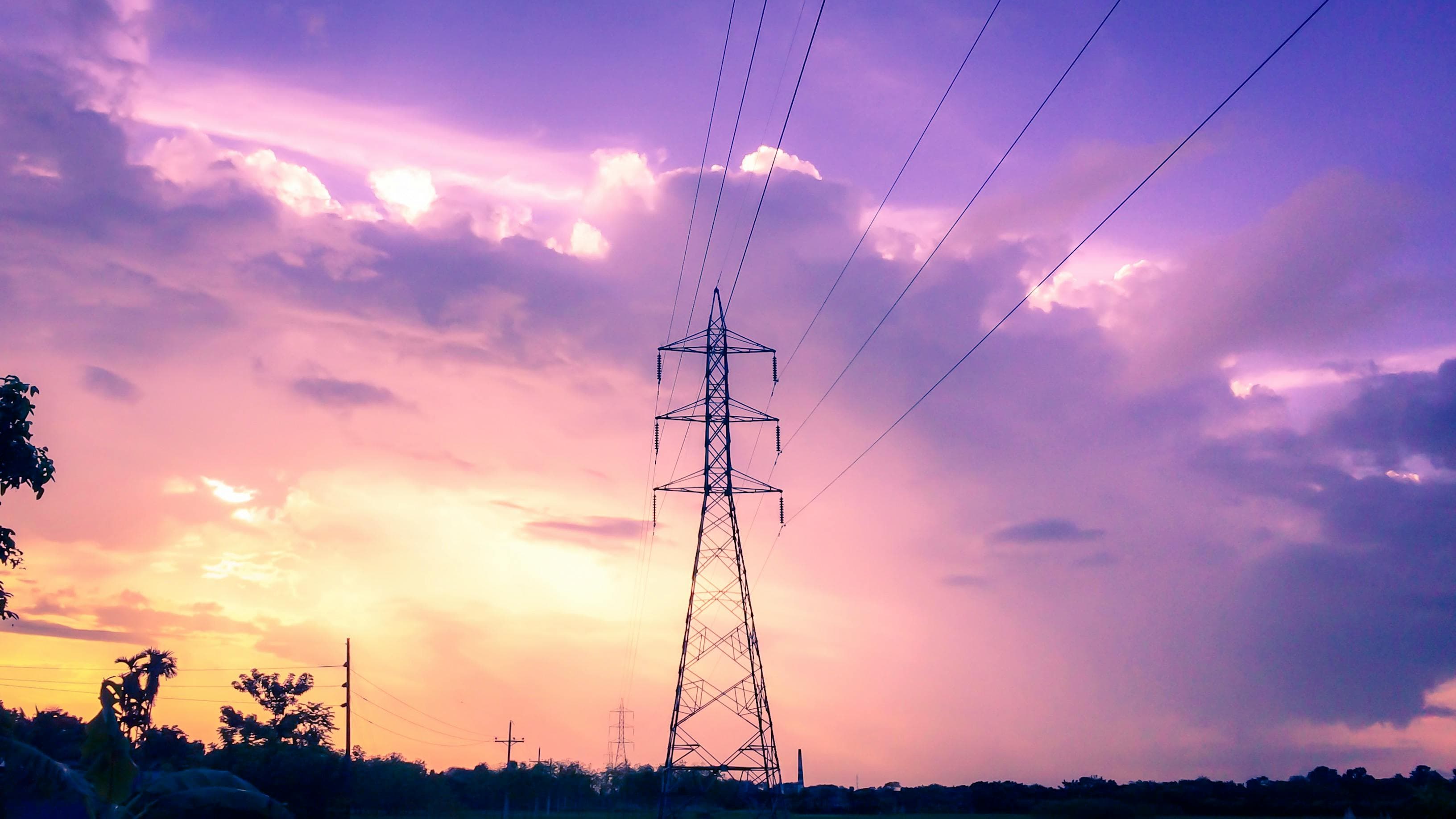 Sähkölinjatorni siluettina eloisaa auringonlaskutaivasta vasten, jossa on violetin ja oranssin sävyjä, hajanaisia pilviä ja kaukana näkyviä puita.