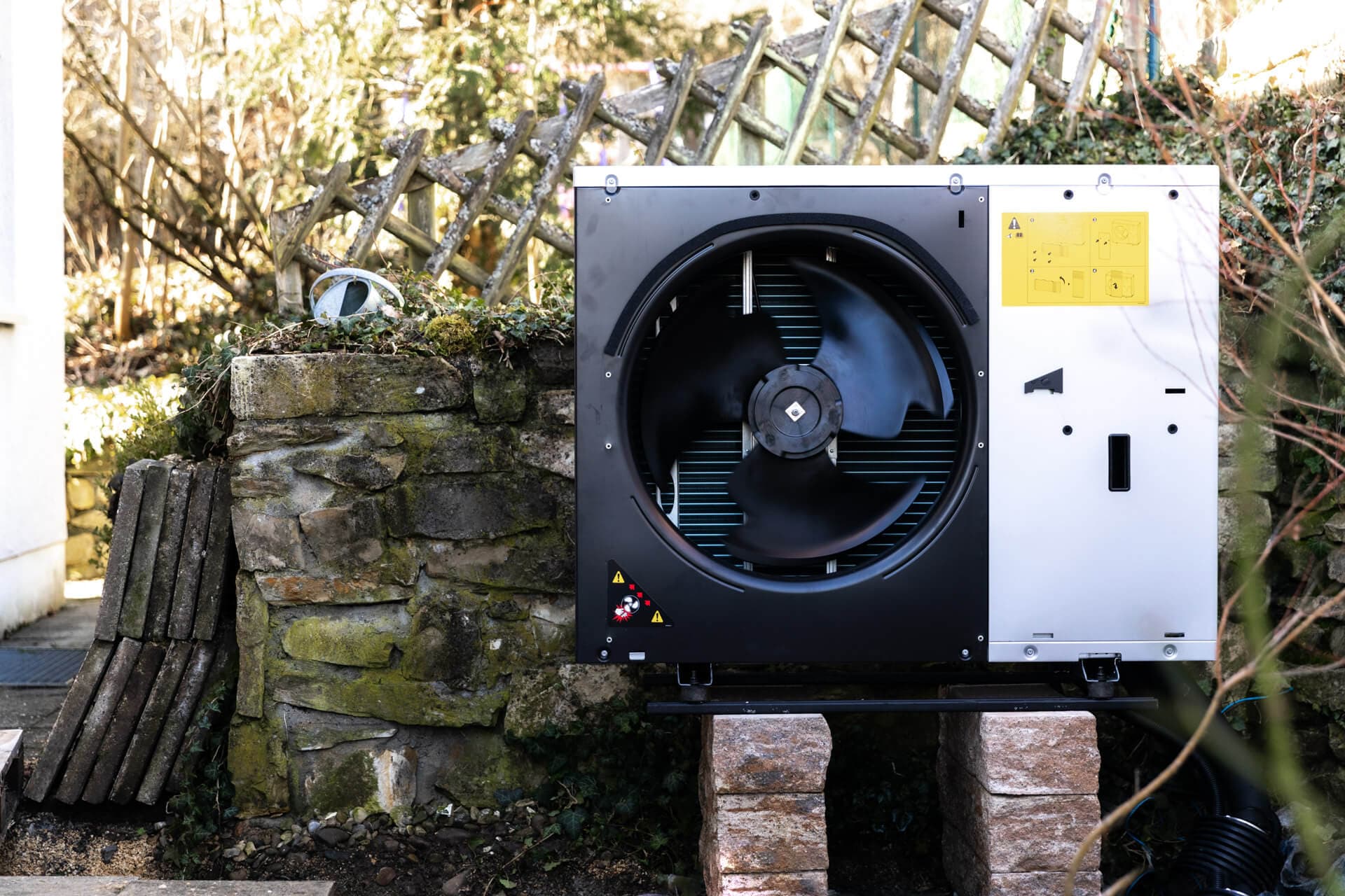 Suuri musta tuuletinyksikkö on asennettu kiviharkkojen päälle ulkona, taustalla rustiikkinen kiviseinä ja puinen säleikkö.