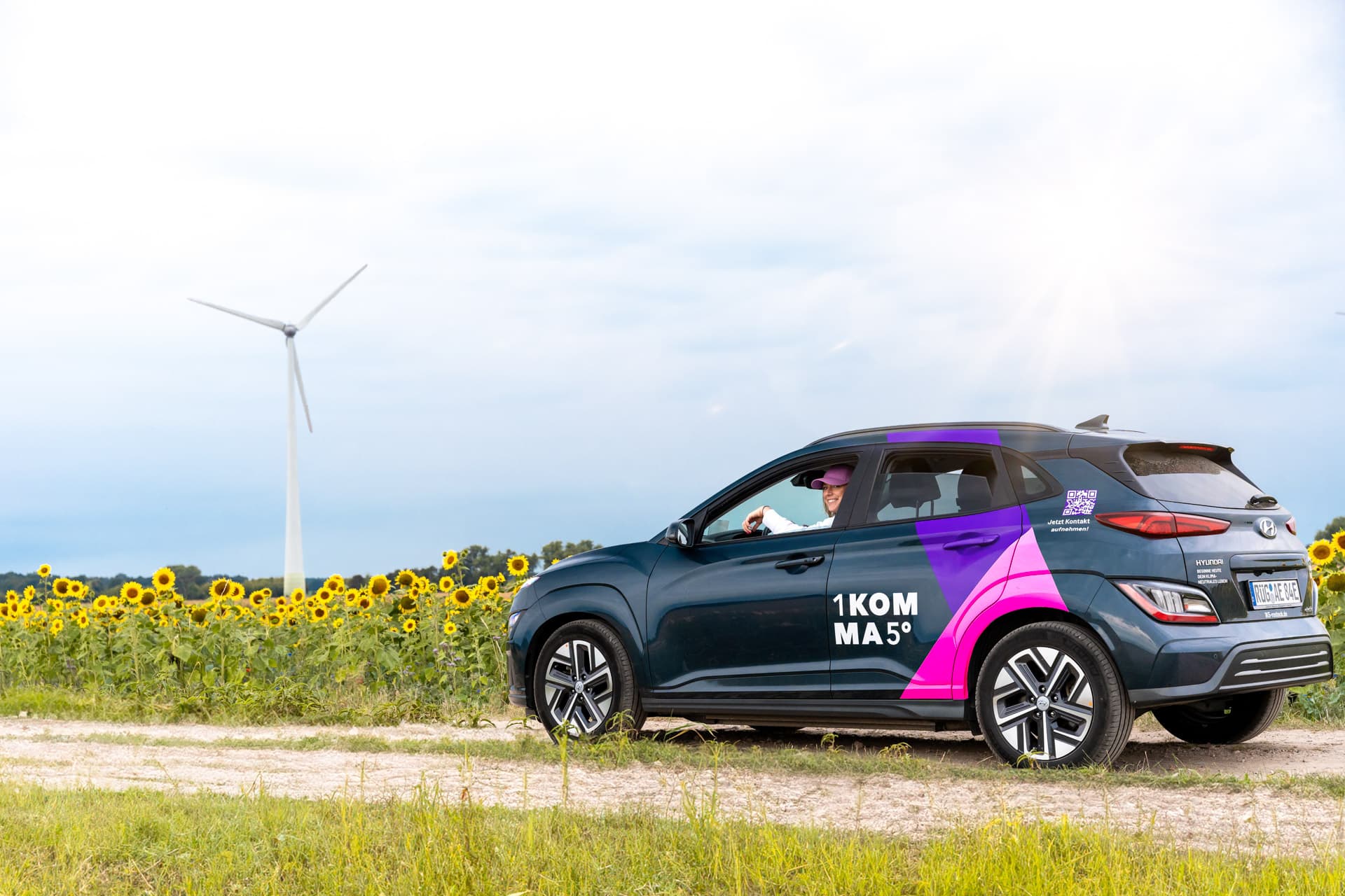 Tumma auto, jossa on pinkki geometrinen kuvio, ajaa soratietä auringonkukkapellon ja tuuliturbiinin vieressä pilvisen taivaan alla.