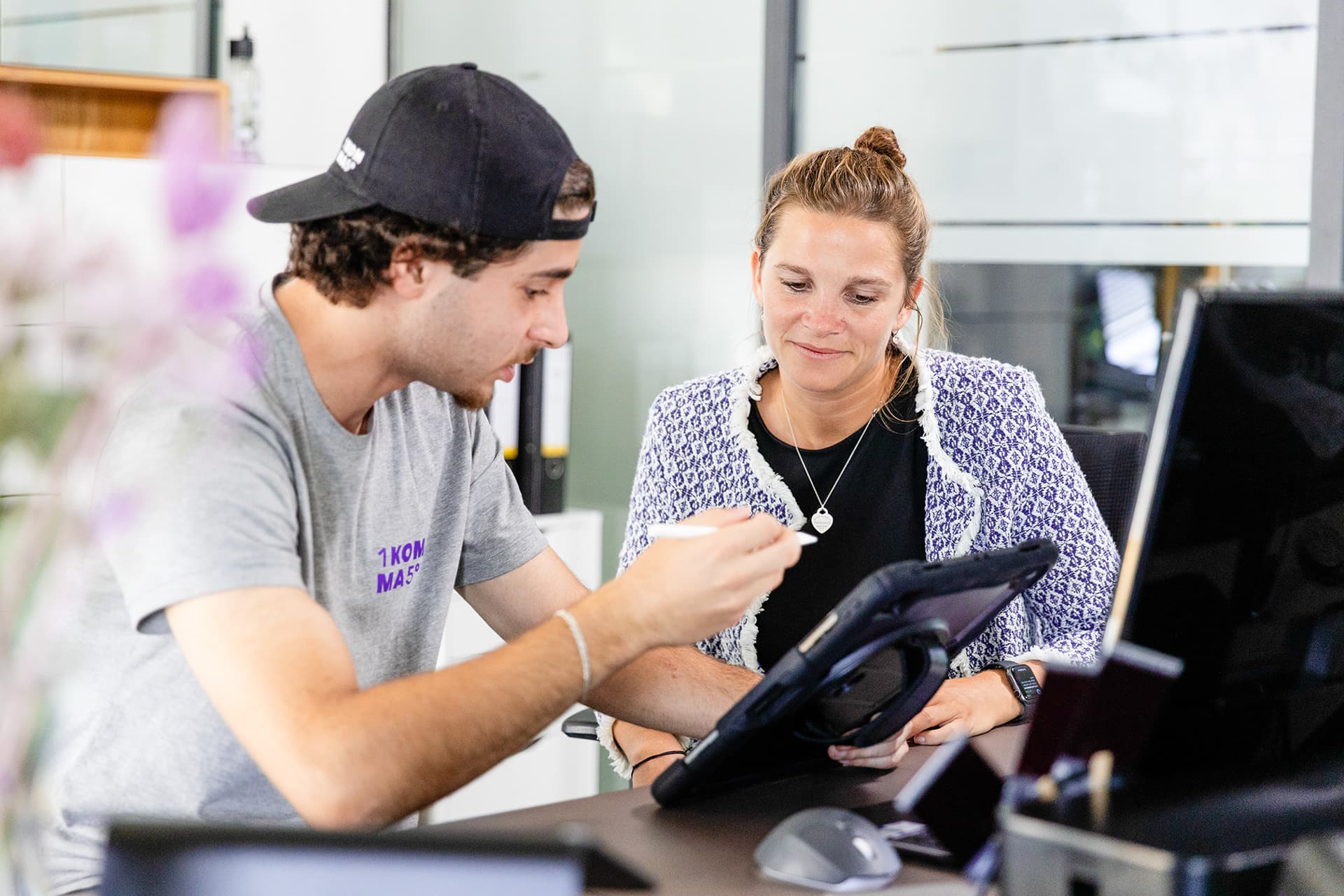 Mies lippis päässään ja nainen keskustelevat jostakin katsoessaan tablettia toimistoympäristössä.