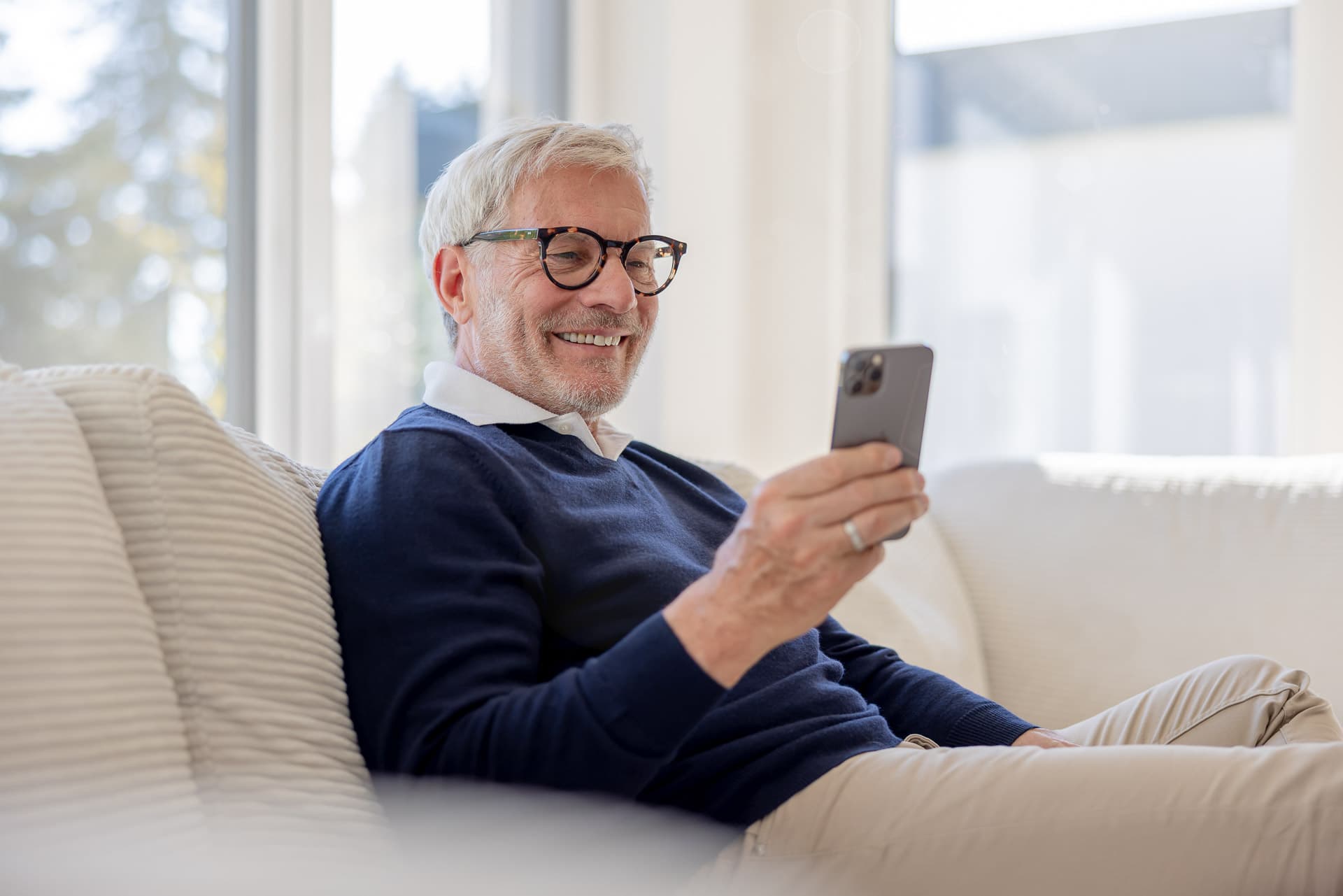 Iäkkäämpi mies silmälaseissa hymyilee älypuhelimelleen istuessaan sohvalla valoisassa huoneessa.