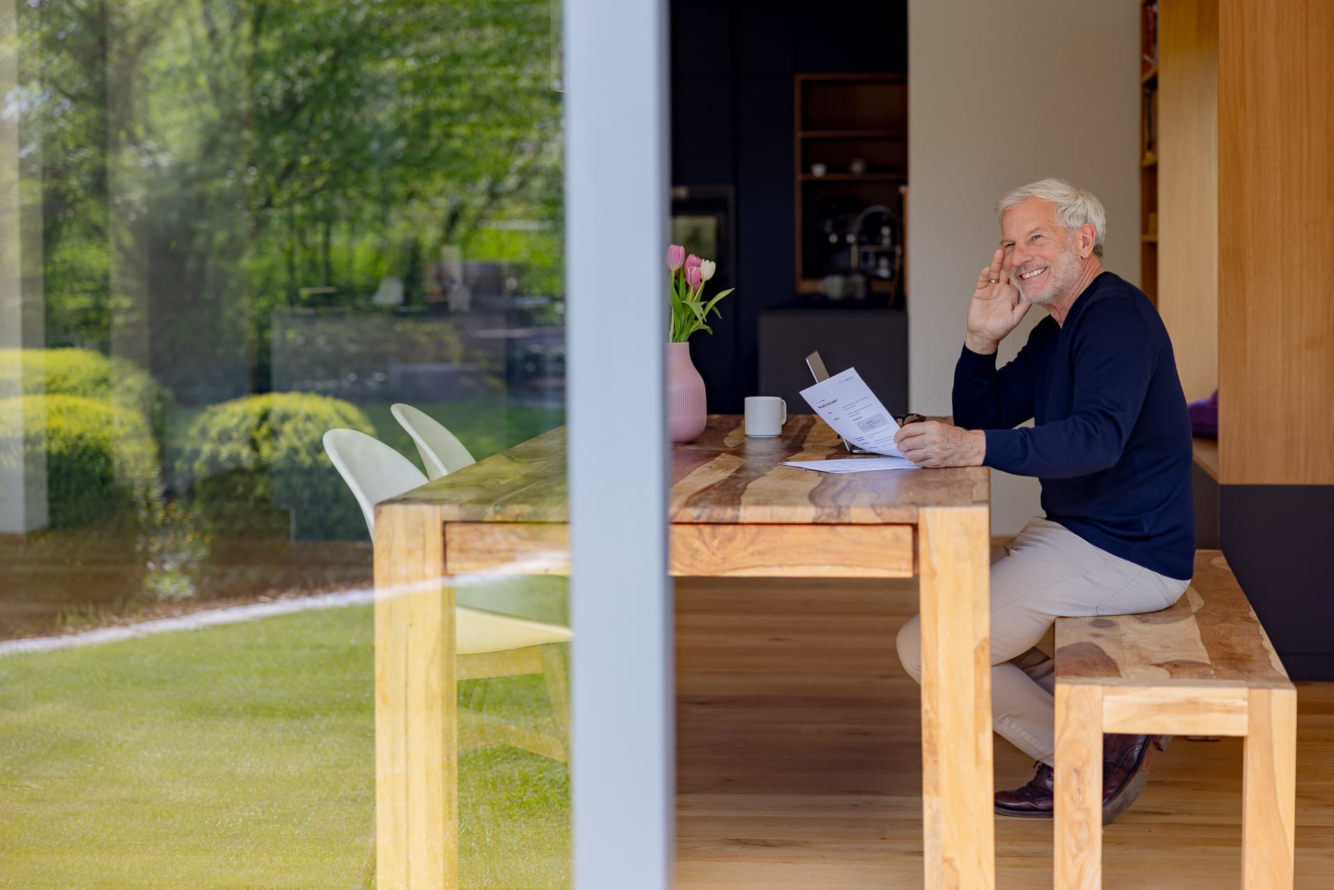 Iäkkäämpi mies hymyilee istuessaan puisessa pöydässä papereiden ja mukin kanssa suuren ikkunan äärellä, josta avautuu näkymä puutarhaan.