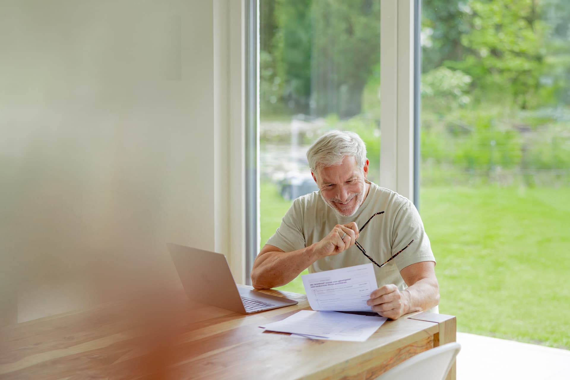 Iäkkäämpi mies hymyilee tarkastellessaan asiakirjoja puupöydän ääressä, jossa on myös kannettava tietokone, suuren ikkunan vieressä, josta avautuu näkymä puutarhaan.