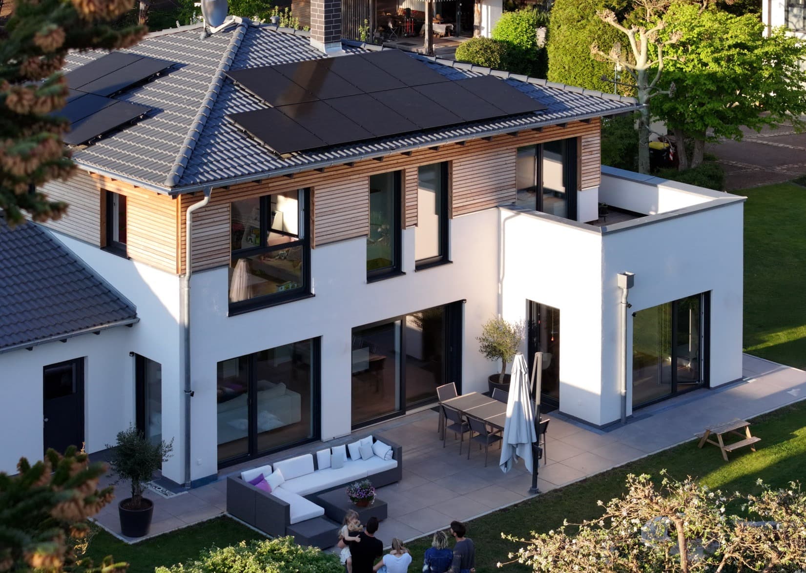 Moderni kaksikerroksinen talo, jossa on aurinkopaneelit, suuret ikkunat ja tilava terassi ulkoistuimilla ja vehreällä ympäristöllä.